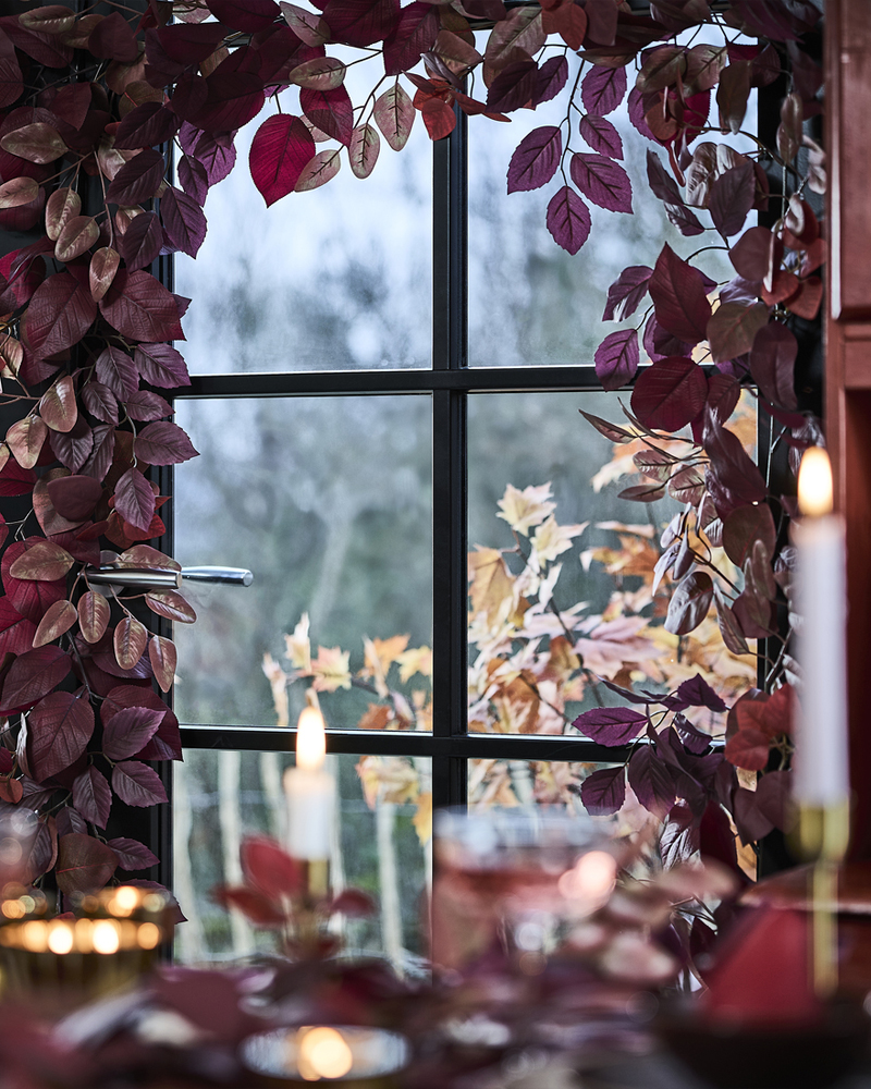 Una ventana de cristal está ricamente decorada con guirnaldas artificiales rojas de SMYCKA, junto a una mesa de comedor con velas encendidas.