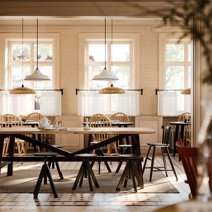 Zona de comedor amplia con mesas de madera, taburetes negros y sillas de barrotes bajo lámparas de luz cálida junto a grandes ventanales con cortinas blancas.