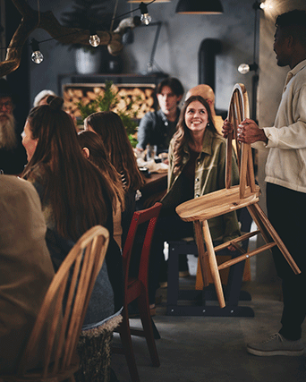 Un hombre acerca una silla de madera clara a una mesa de cena concurrida y festiva, bajo guirnaldas de luces en un interior rústico y cálido.