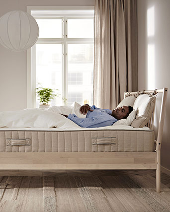 Hombre descansando plácidamente en una cama de madera clara con colchón firme, frente a una ventana luminosa en un ambiente sereno y natural.