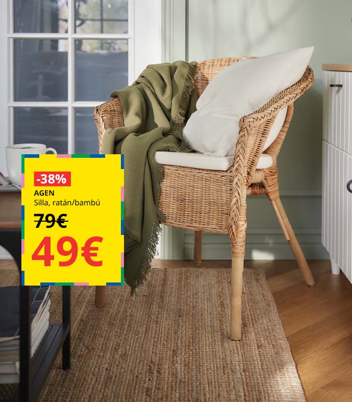 Sillón de ratán con un cojín blanco y una manta verde, junto a una mesa auxiliar y un mueble blanco de salón.